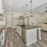 Modern open-concept kitchen with white cabinets, brick backsplash, island with sink, pendant lights, and wood-look flooring. Living area with white sofas, fireplace, and ceiling fan in background.