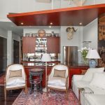 A stylish living room with white seating, patterned rug, Asian-inspired wall art, antique furniture, animal head mounts, and an open kitchen featuring wooden cabinets and stainless steel appliances.
