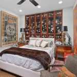 A stylish bedroom with a double bed, brown fur throw, and pillows. Behind the bed is a large decorative panel with Asian-style art. There are ornate side tables with matching lamps and bold modern artwork on the wall.