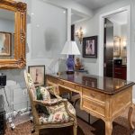 A home office with an ornate wooden desk, floral upholstered chairs, a purple lamp, a large gold-framed mirror, various framed photos and art on the walls, and a printer beside the desk.