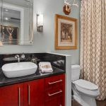 A bathroom with a vessel sink on a dark countertop, red wooden cabinets, a toilet, gold-patterned shower curtain, lamp, wall art, and decorative antlers above the toilet.