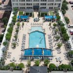 Aerial view of a rooftop swimming pool surrounded by lounge chairs, umbrellas, trees, and people swimming, with a parking lot and tall buildings visible nearby.