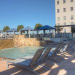 A hotel outdoor pool area with lounge chairs lined up beside the water, stone wall with waterfall, blue umbrellas, and a tall building in the background under a clear blue sky.