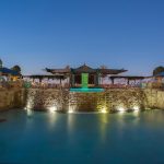 A luxurious outdoor pool with stone walls, soft lighting, and private cabanas on each side at dusk, under a clear blue sky. Trees and lounge chairs surround the pool, creating a tranquil atmosphere.