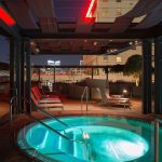 A rooftop hot tub glows at night, surrounded by metal railings, lounge chairs, and decorative trees, with a cityscape visible in the background and a “No Lifeguard on Duty” sign on the stone wall.