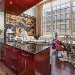 A modern loft apartment with an open kitchen and living area, featuring a large window, eclectic decor, patterned rugs, a kitchen island, and a cozy seating area with various chairs and a sofa.
