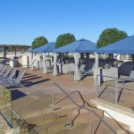 Poolside area with lounge chairs, cabanas with blue canopies, and potted trees under a clear blue sky. Stone steps lead to the pool, surrounded by a metal railing.
