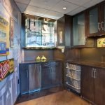 A modern kitchenette with dark cabinets, a mini fridge, a glass candy jar, and wine racks. Vintage soda and candy ads decorate the walls, and glass shelves display memorabilia.