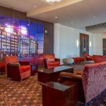 A modern hotel lobby with red chairs, patterned carpet, dark wood tables, and a large mural of a city building on one wall. The space is well-lit and features contemporary design elements.