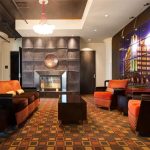 Modern lounge with orange and brown armchairs and sofas, a central coffee table, patterned carpet, a decorative fireplace, and a brightly lit wall featuring cityscape images.