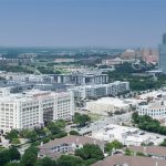 Aerial view of a cityscape with modern and historical buildings, green trees, and roads. A large white building labeled "Montgomery Plaza" stands out in the foreground, with high-rise buildings in the distant background.