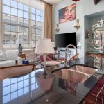 Modern loft living room with large windows, a white sectional sofa, a TV, eclectic artwork, ornate mirror, and a glossy kitchen island with a sink in the foreground. Natural light fills the stylish, open space.
