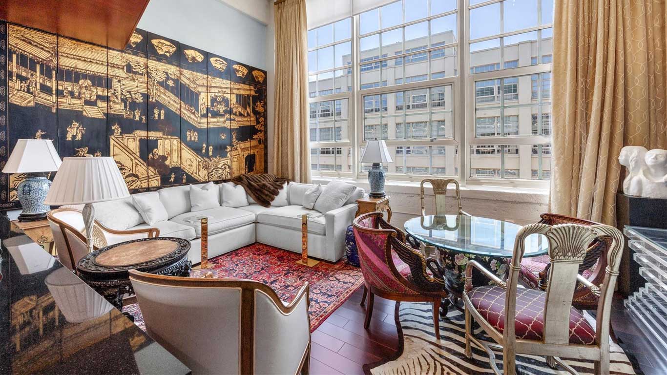 A stylish living room with a white sectional sofa, eclectic chairs around a glass table, a vibrant rug, zebra-patterned rug, large windows with city views, and a large black and gold mural on one wall.