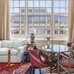 A stylish living room with a white sectional sofa, glass dining table with ornate chairs, red patterned rug, large windows with beige curtains, and eclectic art and decor throughout.