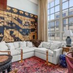A bright living room with a large window, white sectional sofa, ornate gold and black wall art, vintage tables, traditional lamps, and a red patterned rug on the floor.