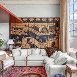 A bright, stylish living room with a large sectional sofa, ornate chairs, table lamps, a red Persian rug, and a decorative wall mural. Large windows let in natural light, revealing an urban city view outside.