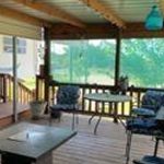 A covered wooden porch with a metal roof features cushioned chairs, a small table, and a potted plant. Large windows reveal green trees and grass outside, creating a cozy, inviting outdoor seating area.