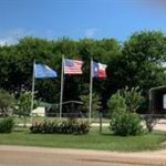 Three flags—the Oklahoma, U.S., and Texas flags—fly on flagpoles in front of a fenced property with trees, a driveway, and a metal building under a partly cloudy sky.