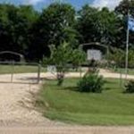 A gravel driveway leads to a gated entrance with two flagpoles, one flying the American flag, surrounded by green grass, shrubs, and trees under a partly cloudy sky.