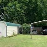 A white shed with a green roof sits next to a carport sheltering a black pickup truck, set in a grassy yard with trees and shrubs in the background.