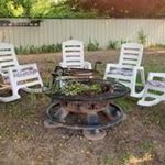 Six white rocking chairs with cushions are arranged in a circle around a metal fire pit in a backyard with a dirt ground, trees, and a white fence in the background.