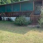 A small house with a covered front porch, wooden railings, and steps, surrounded by greenery and a grassy yard. Trees and bushes are visible near the house.