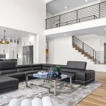 Modern open-concept living room with a large black sectional sofa, glass coffee table, kitchen with island, and a black grand piano. Light wood floors and a staircase lead to an upper loft with a metal railing.