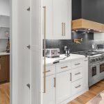 Modern kitchen with white cabinets, stainless steel appliances, a gas stove, chevron-patterned backsplash, wood flooring, and a separate pantry area with brown cabinets, shelves, and a microwave.