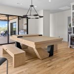 Modern dining area with light wood table and bench, black chair, and stylish black chandelier. Glass doors lead to a patio with outdoor seating. White cabinets, a coffee machine, and a wine fridge are visible in the background.