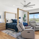 A modern bedroom with a wooden canopy bed, blue bedding, and two beige recliners facing the bed. Large windows bring in natural light, and there are plants and wooden furniture on a light wood floor.