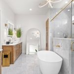 Modern bathroom with white and wood decor, featuring a freestanding bathtub, glass shower, large mirror, double sinks with wooden cabinets, wall sconces, and plants for decoration. Marble floors and bright lighting.