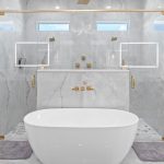 Modern bathroom with a freestanding white tub, marble walls, dual glass shower with gold fixtures, two mirrors, and wooden cabinetry on the left. Two gray bath mats are placed on the floor.