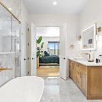Modern bathroom with marble floors, a freestanding bathtub, glass shower, wood vanities with white countertops, gold fixtures, and a large mirror; double doors open to a bright bedroom with plants.
