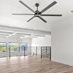 A modern loft space with light wood flooring, white walls, a black ceiling fan, and a metal railing overlooking large windows that let in natural light and offer a view of greenery outside.