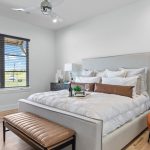 A modern bedroom with a large bed, white bedding, a tan bench at the foot, an orange armchair, wooden floors, a large window with blinds, and minimal decor.