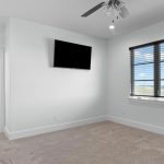 A bright, carpeted room with an exercise bike, wall-mounted TV, ceiling fan, open door to a bathroom, and a window with blinds showing a scenic outdoor view.