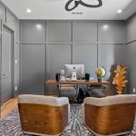 Modern home office with gray paneled walls, a large wooden desk facing two mid-century chairs, a Mac computer, large window with blinds, shelves with books and decor, and a patterned rug on light wood flooring.