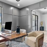 A modern home office with a wooden desk, computer, microphone, two beige chairs, large window with blinds, gray paneled walls, a patterned rug, and a tall green plant in the corner.