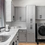 A modern laundry room with gray cabinets, a stainless steel sink, a black faucet, and a washer and dryer set. Large window with dark blinds lets in natural light. White countertops and light wood flooring complete the space.