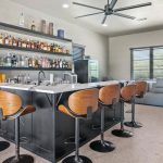 Modern home bar with black countertop, wood-backed barstools, shelves stocked with bottles and glassware, a sink, and a lounge area with a sofa and large windows letting in natural light.