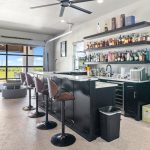 Modern home bar with stools, shelves stocked with liquor bottles, a counter with a sink, mini-fridge, glass-door fridge, and large window showing an outdoor view with blue sky and greenery.