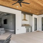 Covered patio with wood ceiling, ceiling fans, and outdoor grill. Sliding doors open to a modern indoor living space with bar seating and a television. Adjacent glass doors reveal a dining area and windows with greenery outside.