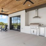 A covered patio with a wood ceiling, ceiling fans, a built-in stainless steel grill, and black-framed glass doors. Four Adirondack chairs face a grassy field and blue sky in the background.
