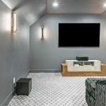 A modern home theater room with gray walls, patterned carpet, wall-mounted TV, light fixtures, a wooden media console, speakers, and a plush dark sofa in the foreground.