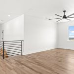 A spacious, modern room with light wood floors, white walls, a black ceiling fan, a large window with a scenic view, and a black metal railing along a staircase.