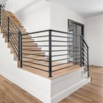 A modern, open hallway with light wood floors, a central staircase with black metal railings, white walls, large windows, and glass doors letting in natural light and offering a view of the outdoors.