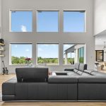 A modern living room with a black sectional sofa, large windows showing a scenic outdoor view, a mounted flat-screen TV above a fireplace, and an open kitchen and dining area in the background.