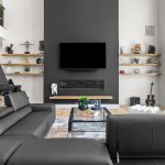Modern living room with black couches, a large TV mounted on a dark accent wall, open shelves with decor, a glass coffee table, a grand piano, a guitar, and big windows with views of greenery outside.