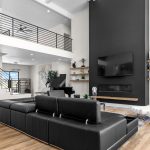 Modern living room with black leather sectional sofa, white armchairs, large black accent wall with mounted TV, fireplace, open staircase, wooden floors, and minimalist decor on shelves. Bright and spacious with high ceilings.