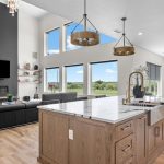 Modern open-concept living room and kitchen with large windows, light wood floors, a marble island, and a black sectional sofa. Scenic green landscape visible through the windows.